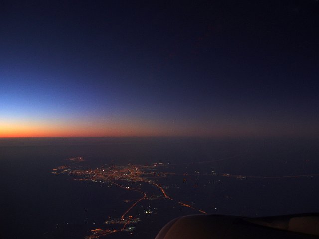 View from an airplane window of the landscape below at dusk with colorful sunset on the horizon and twinkling lights of a town