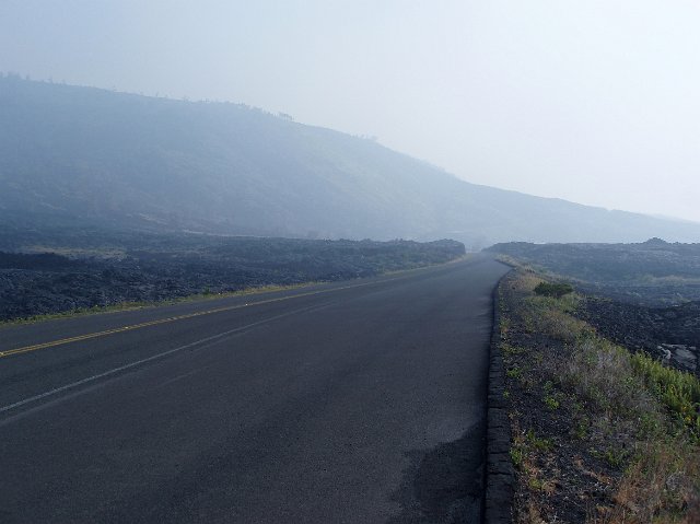 Empty country road through the meadow with burnt grass after the fire and barely seen hill through the smoky air. Dystopian landscape in greyish colors