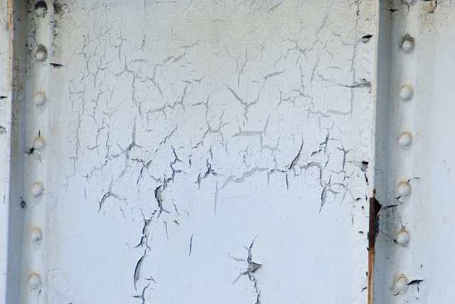 Weathered metal panel with rivets and cracked peeling white paint showing signs of corrosion and rust in a full frame background